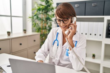 Genç, güzel, İspanyol bir kadın. Laptop kullanıyor. Klinikte telefonla konuşuyor.