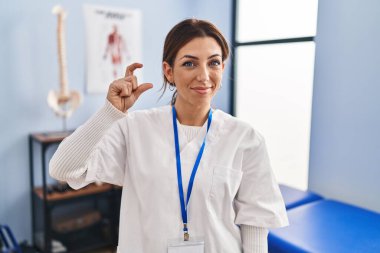 Young brunette woman working at pain recovery clinic smiling and confident gesturing with hand doing small size sign with fingers looking and the camera. measure concept. 