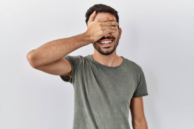 Young hispanic man with beard wearing casual t shirt over white background smiling and laughing with hand on face covering eyes for surprise. blind concept. 