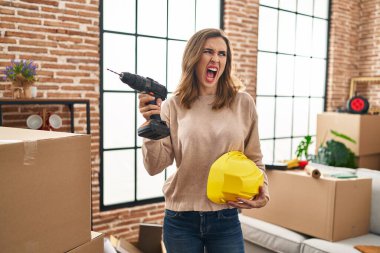 Young woman holding drill at new home angry and mad screaming frustrated and furious, shouting with anger. rage and aggressive concept. 