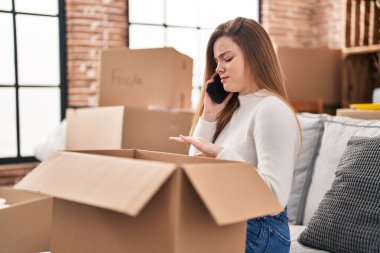 Young blonde woman unboxing package talking on the smartphone at new home