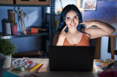 Young modern girl with blue hair sitting at art studio with laptop at night smiling cheerful showing and pointing with fingers teeth and mouth. dental health concept. 
