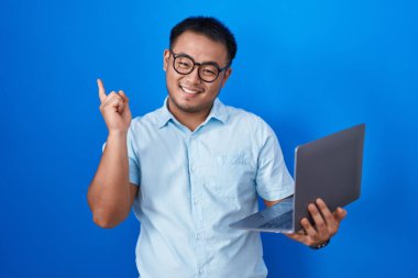 Chinese young man using computer laptop with a big smile on face, pointing with hand finger to the side looking at the camera. 