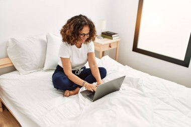 Orta yaşlı İspanyol kadın yatak odasında dizüstü bilgisayar kullanarak gülümsüyor.