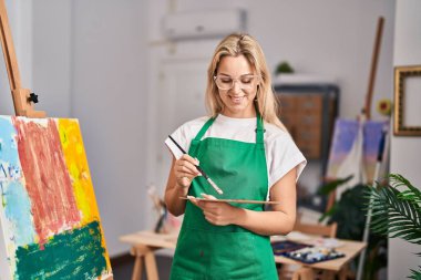 Sanat stüdyosunda kendine güvenen genç sarışın sanatçı gülümsüyor.