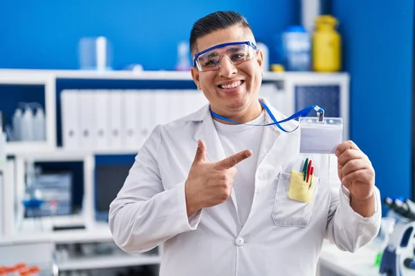 Hispanic young man working at scientist laboratory holding id card smiling happy pointing with hand and finger