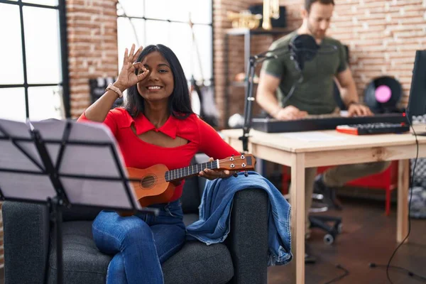 Young indian woman playing ukulele at music studio smiling happy doing ok sign with hand on eye looking through fingers