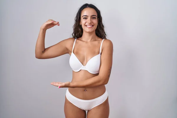 Young hispanic woman wearing white lingerie gesturing with hands showing big and large size sign, measure symbol. smiling looking at the camera. measuring concept.