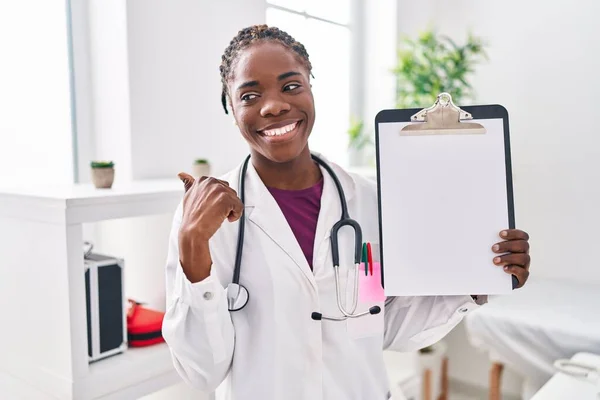 Beautiful black doctor woman holding clipboard pointing thumb up to the side smiling happy with open mouth
