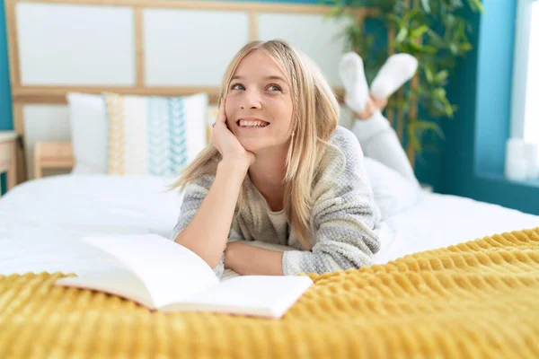 Young blonde woman reading book lying on bed at bedroom