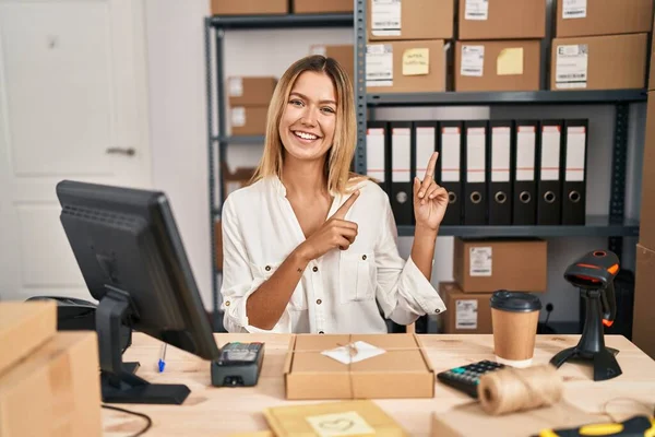 Young blonde woman working at small business ecommerce smiling and looking at the camera pointing with two hands and fingers to the side.