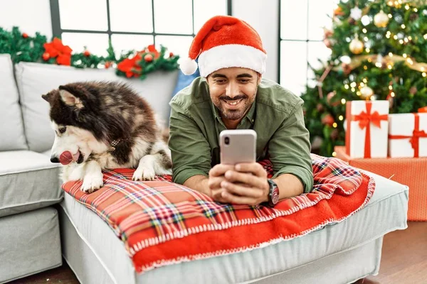 Young Hispanic Man Using Smartphone Lying Sofa Dog Christmas Tree — Stockfoto
