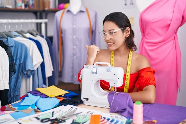 Hispanic young woman dressmaker designer using sewing machine smiling with happy face looking and pointing to the side with thumb up.