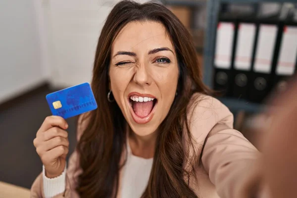 Young brunette woman working at small business ecommerce holding credit card winking looking at the camera with sexy expression, cheerful and happy face.