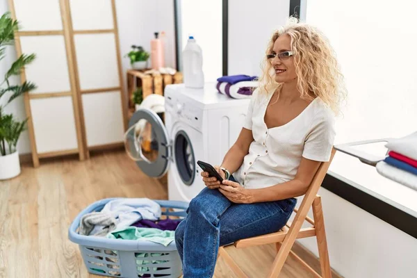 Medelålders Blond Kvinna Med Smartphone Väntar Tvättmaskin Förråd — Stockfoto