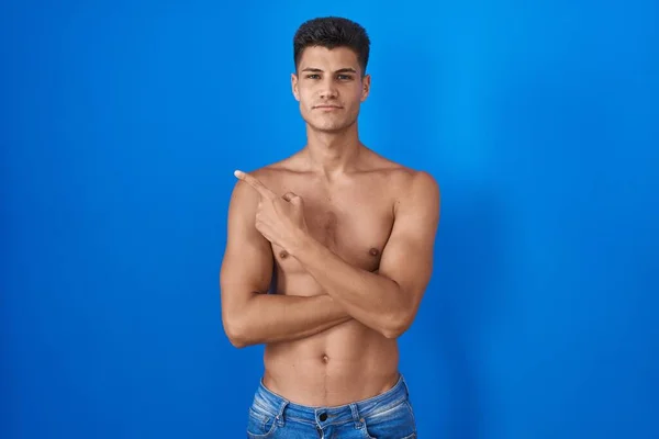 Young hispanic man standing shirtless over blue background pointing with hand finger to the side showing advertisement, serious and calm face