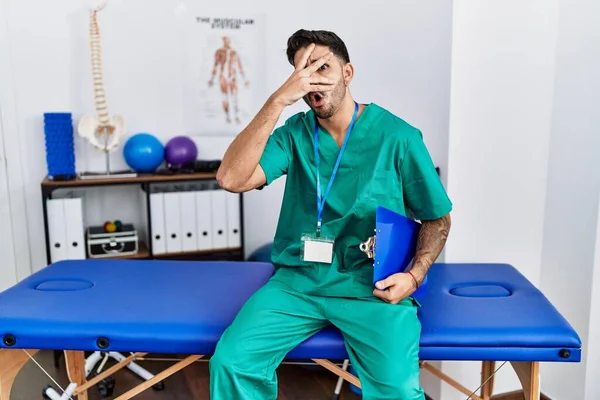 Young physiotherapist man working at pain recovery clinic peeking in shock covering face and eyes with hand, looking through fingers with embarrassed expression.