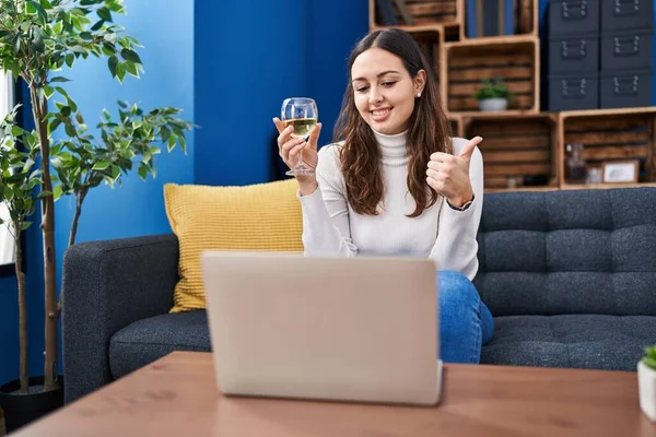 Young hispanic woman doing video call drinking white wine smiling happy and positive, thumb up doing excellent and approval sign