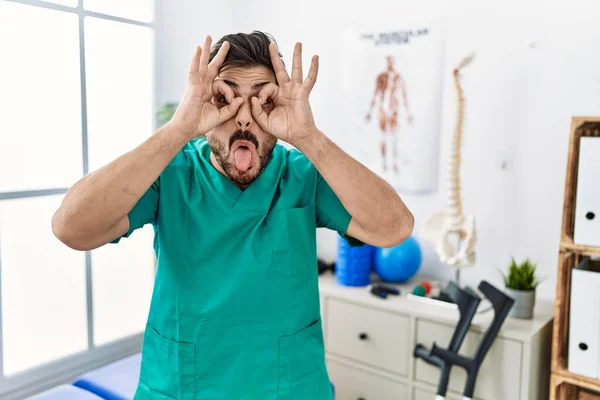 Young man with beard working at pain recovery clinic doing ok gesture like binoculars sticking tongue out, eyes looking through fingers. crazy expression.