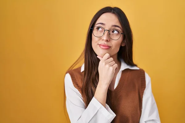 Young Brunette Woman Standing Yellow Background Wearing Glasses Hand Chin — Zdjęcie stockowe