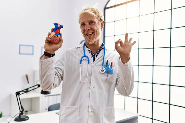 Młody Blondyn Noszący Mundur Lekarza Trzymający Serce Klinice Robi Znak — Zdjęcie stockowe