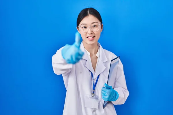 Chinese young woman working at scientist laboratory approving doing positive gesture with hand, thumbs up smiling and happy for success. winner gesture.