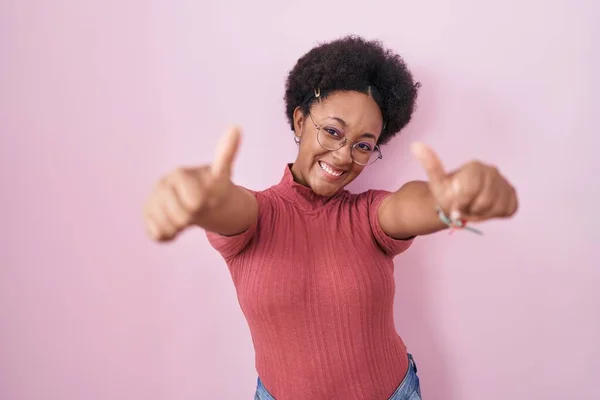 ピンクの背景の上に立って巻き毛を持つ美しいアフリカの女性は 手で肯定的なジェスチャーを行うことを承認します 親指アップ笑顔と成功のために幸せ 勝者のジェスチャー — ストック写真