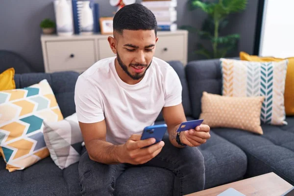 年轻的拉丁男人坐在家里沙发上 用智能手机和信用卡 — 图库照片