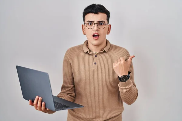 Non binary person using computer laptop surprised pointing with hand finger to the side, open mouth amazed expression.