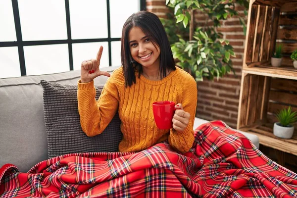 Young hispanic woman sitting on the sofa drinking a coffee at home smiling and confident gesturing with hand doing small size sign with fingers looking and the camera. measure concept.