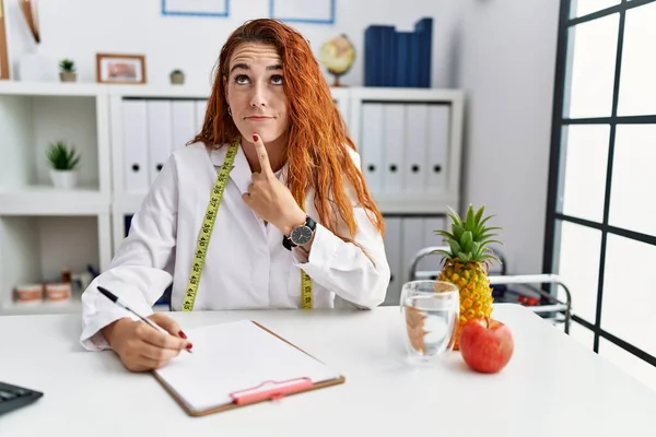 stock image Young redhead woman nutritionist doctor at the clinic thinking concentrated about doubt with finger on chin and looking up wondering 