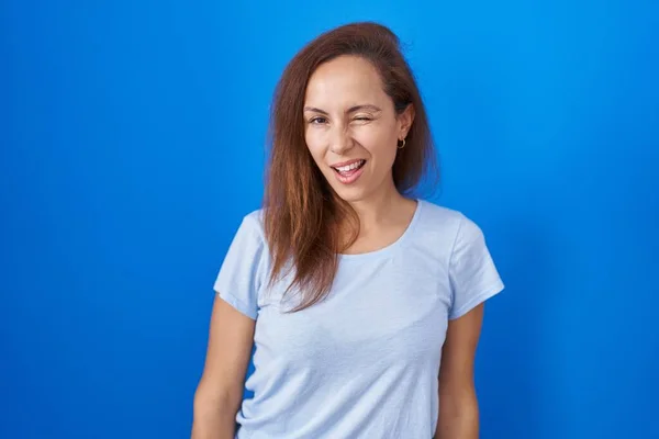 Brunette woman standing over blue background winking looking at the camera with sexy expression, cheerful and happy face.
