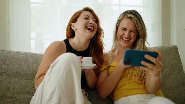 Two women drinking coffee watching video on smartphone at home