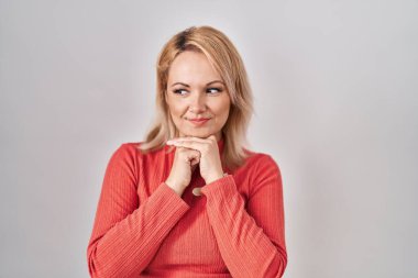 Blonde woman standing over isolated background laughing nervous and excited with hands on chin looking to the side 