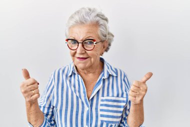 Beyaz arka plan başarı işaretinin üzerinde duran gri saçlı yaşlı bir kadın elleriyle olumlu bir jest yapıyor, gülümsüyor ve mutlu. Neşeli ifade ve kazanan jest. 