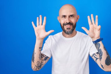 Hispanic man with tattoos standing over blue background showing and pointing up with fingers number nine while smiling confident and happy. 