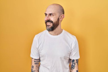Young hispanic man with tattoos standing over yellow background looking away to side with smile on face, natural expression. laughing confident. 