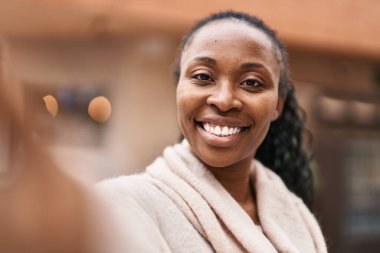 Afro-Amerikalı kadın gülümsüyor. Sokakta akıllı telefonun yanında özçekim yapıyor.