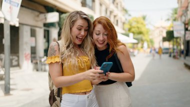 Two women standing together using smartphone at street