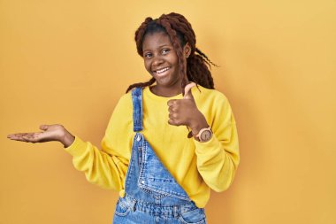 African woman standing over yellow background showing palm hand and doing ok gesture with thumbs up, smiling happy and cheerful 