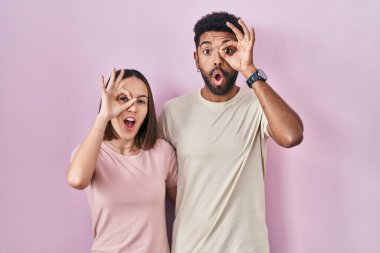 Young hispanic couple together over pink background doing ok gesture shocked with surprised face, eye looking through fingers. unbelieving expression. 