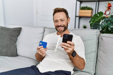 Orta yaşlı İspanyol adam evde akıllı telefon ve kredi kartı kullanıyor.