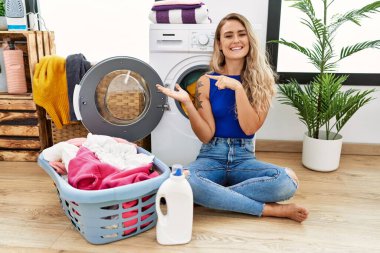 Young beautiful woman doing laundry sitting by wicker basket amazed and smiling to the camera while presenting with hand and pointing with finger. 