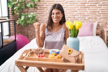 Brunette young woman eating breakfast sitting on the bed screaming proud, celebrating victory and success very excited with raised arms 