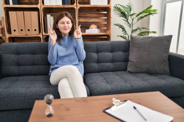 Young brunette woman at consultation office gesturing finger crossed smiling with hope and eyes closed. luck and superstitious concept. 