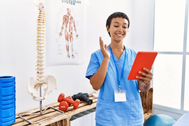 Fizyoterapi kliniğinde fizyoterapist üniforması giyen genç İspanyol kadın görüntülü görüşme yapıyor.
