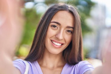 Genç, güzel, İspanyol bir kadın gülümsüyor. Parkta kamerayla selfie çekiyor.