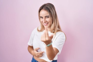 Young blonde woman standing over pink background beckoning come here gesture with hand inviting welcoming happy and smiling 