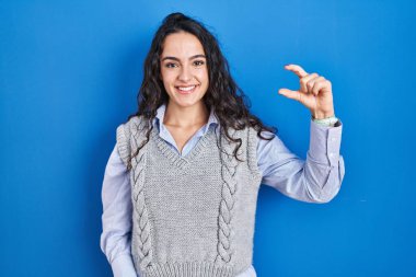 Genç esmer kadın mavi arka planda duruyor gülümsüyor ve el hareketleriyle küçük boy işareti yapıyor ve kameraya bakıyor. ölçüm kavramı. 