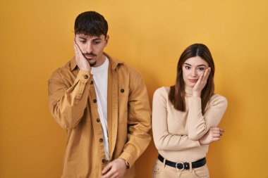 Young hispanic couple standing over yellow background thinking looking tired and bored with depression problems with crossed arms. 